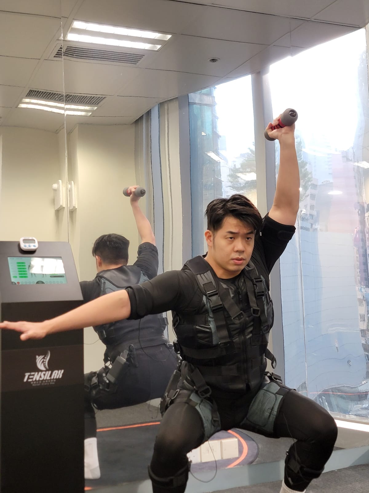 <img src="A male doing a a legs exercise with one arm overhead press with a dumbbell, wearing electric suits stimulating="男子示範單臂使用啞鈴，並身穿電刺激衣服作刺激下肢訓練">